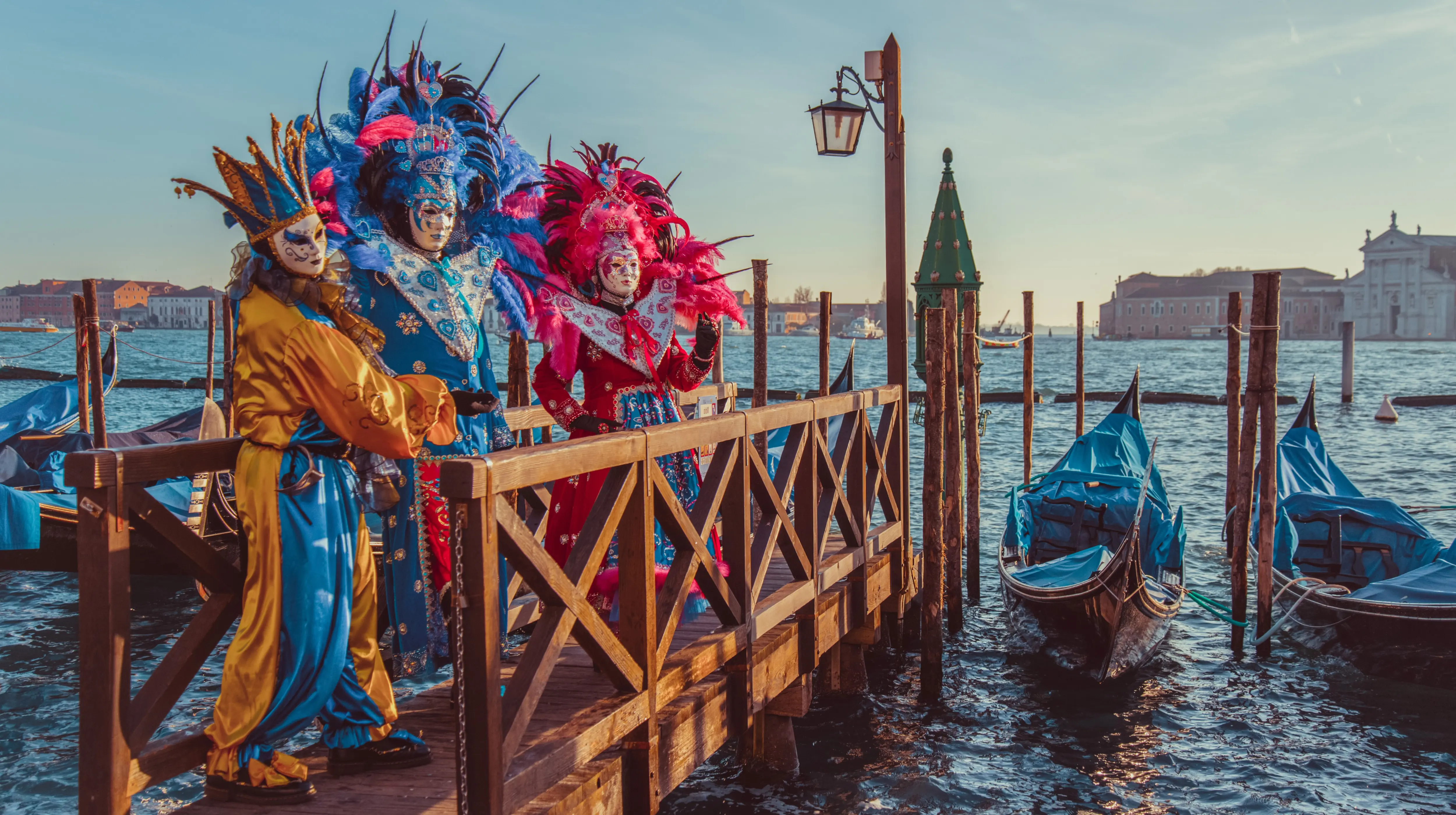 Costumed participants wearing traditional Volto masks Venice Carnival standing wooden pier beside gondolas along Venetian Lagoon Venice Italy