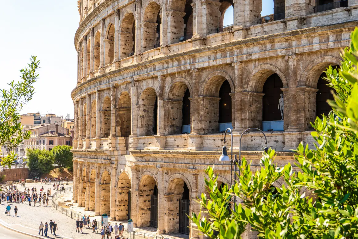 The Colosseum exterior in Rome showing the massive arches and travertine stone facade with visitors below on a warm sunny day
