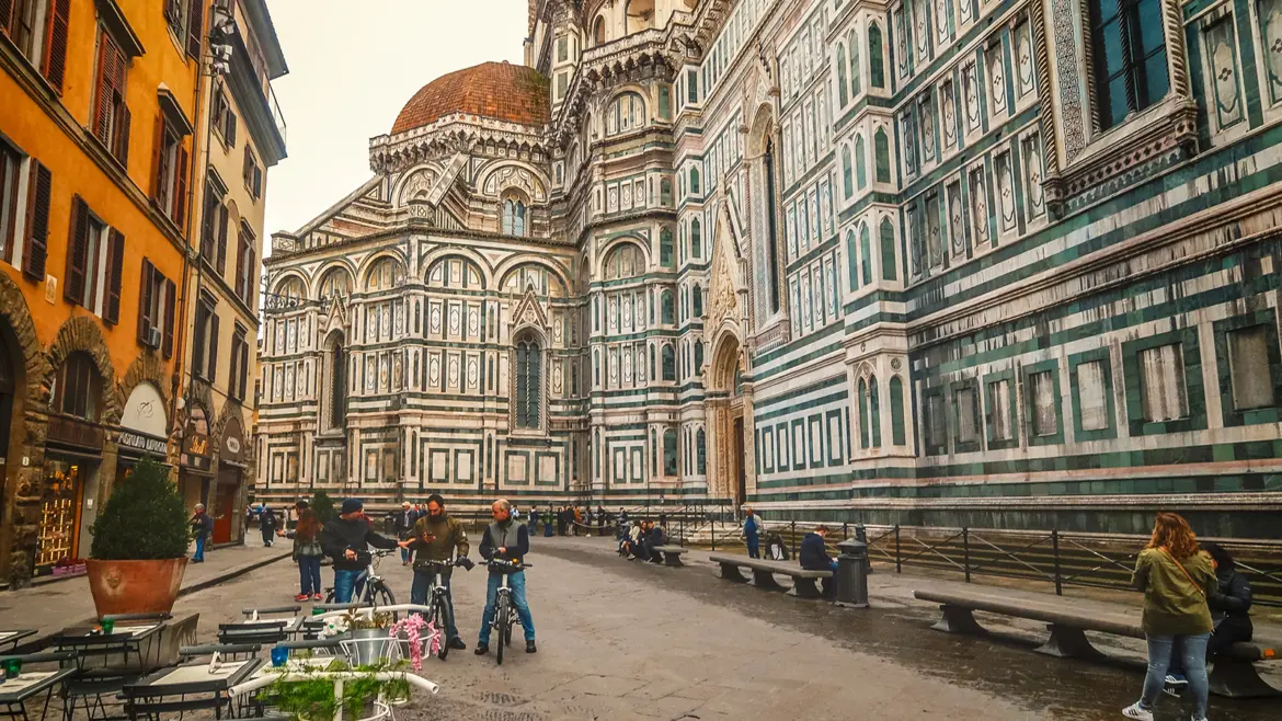 Side exterior of Florence Cathedral in Piazza del Duomo highlighting the intricate green white and pink marble patterns and Gothic architecture as daily life unfolds around the historic landmark