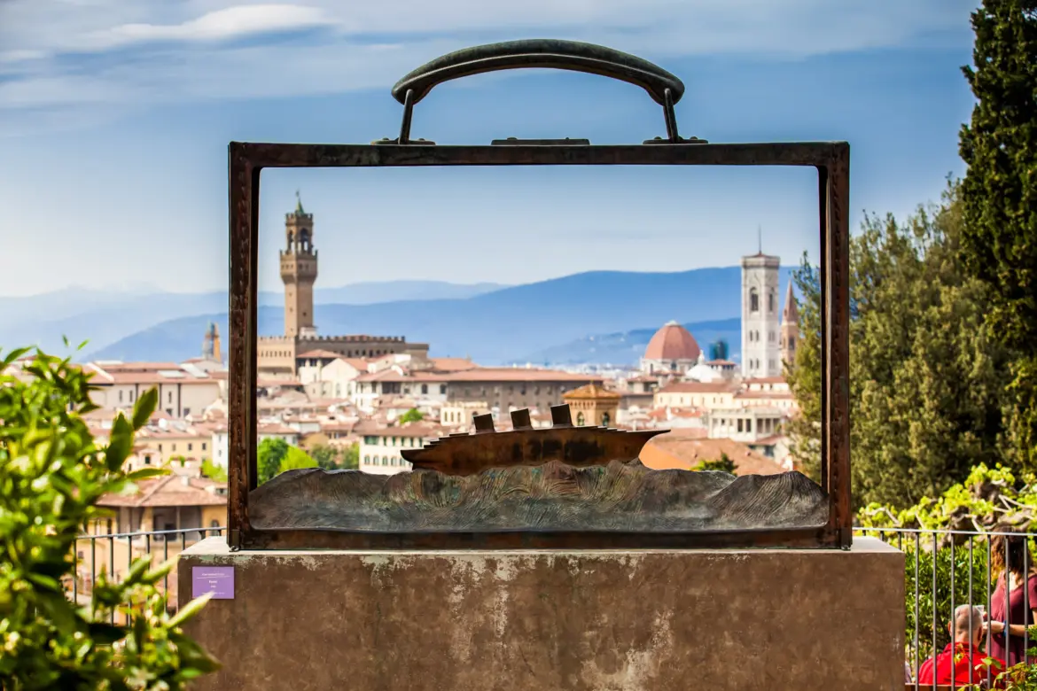 Jean-Michel Folon’s bronze sculpture Partir at Giardino delle Rose in Florence, its open suitcase-shaped frame revealing the Palazzo Vecchio and Duomo in the cityscape beyond