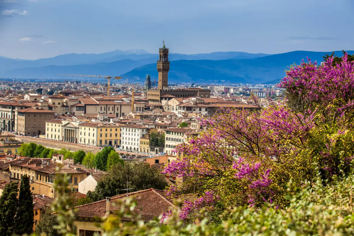 Panoramic view of the Florence skyline from Giardino delle Rose, with the Palazzo Vecchio tower rising above terracotta rooftops and purple spring blossoms framing the Tuscan hills beyond