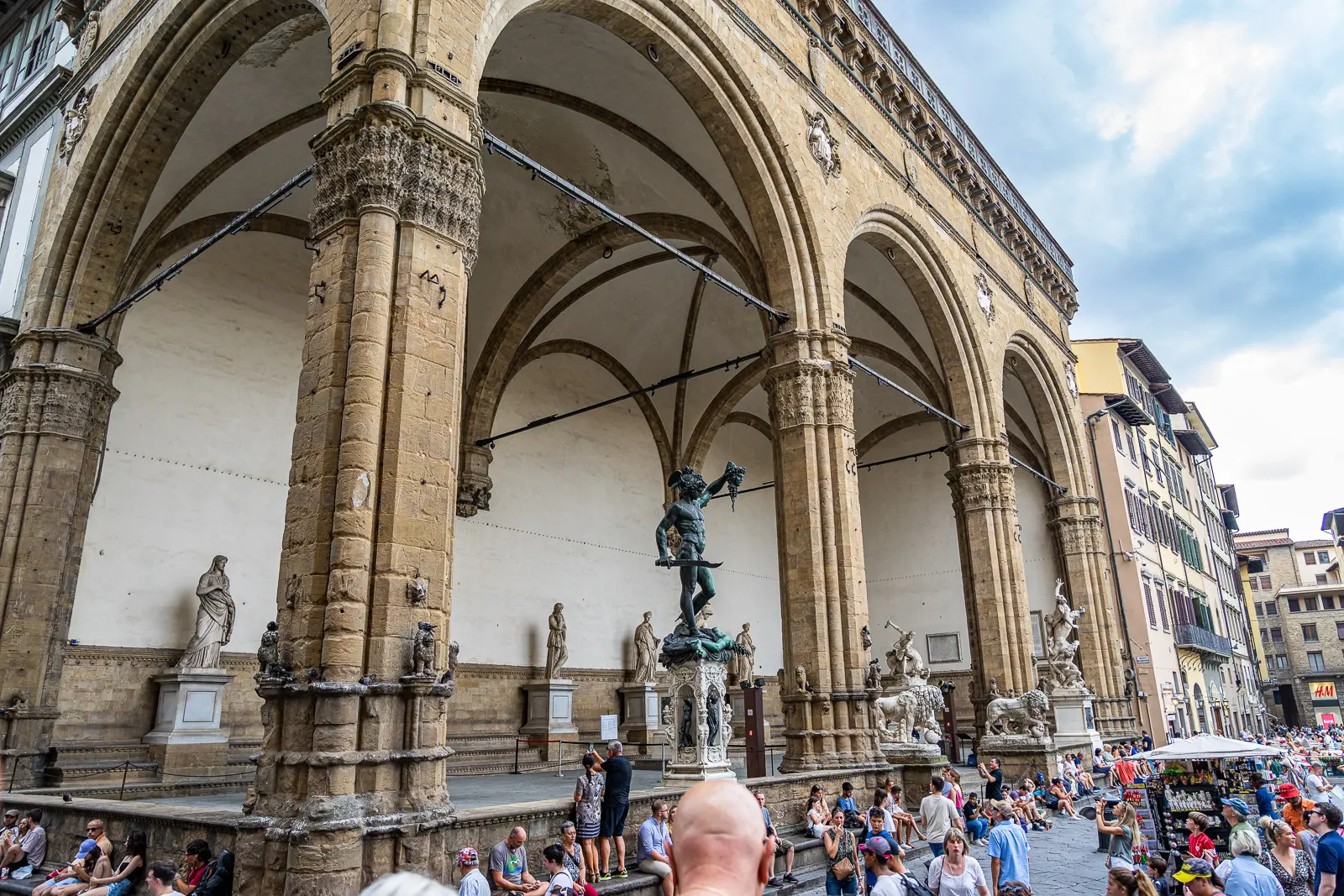 Crowds gathered at the Loggia dei Lanzi in Florence with Cellini Perseus sculpture visible through Gothic arches and sculptures lining the gallery