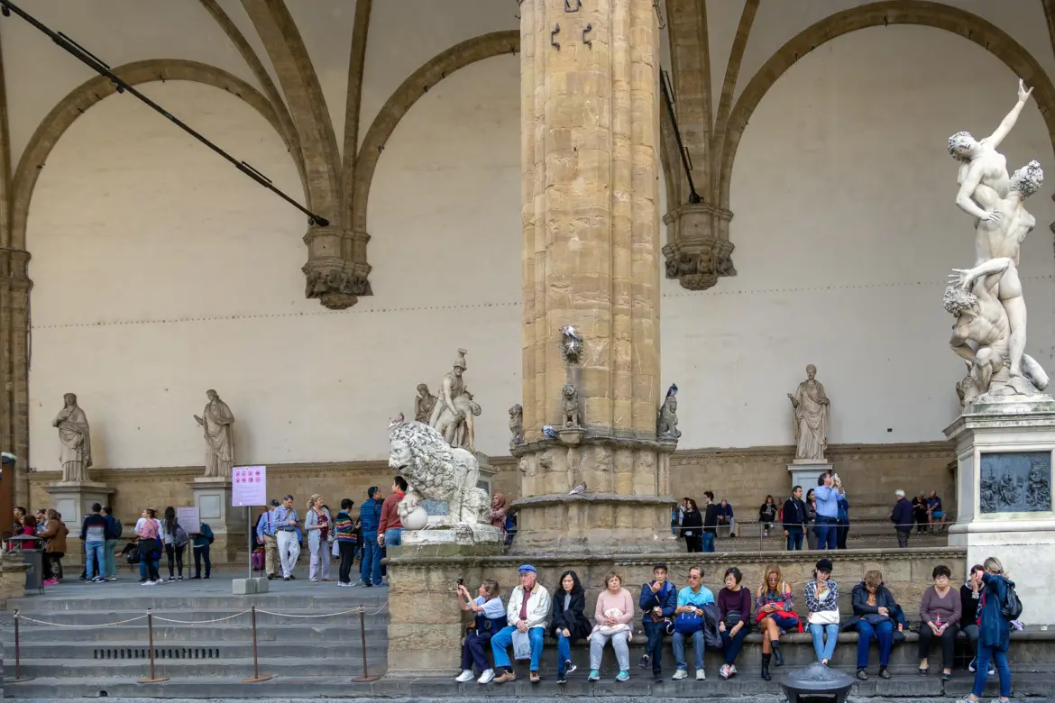 Visitors sitting on the steps of the Loggia dei Lanzi in Florence with Renaissance sculptures visible beneath the arches behind them