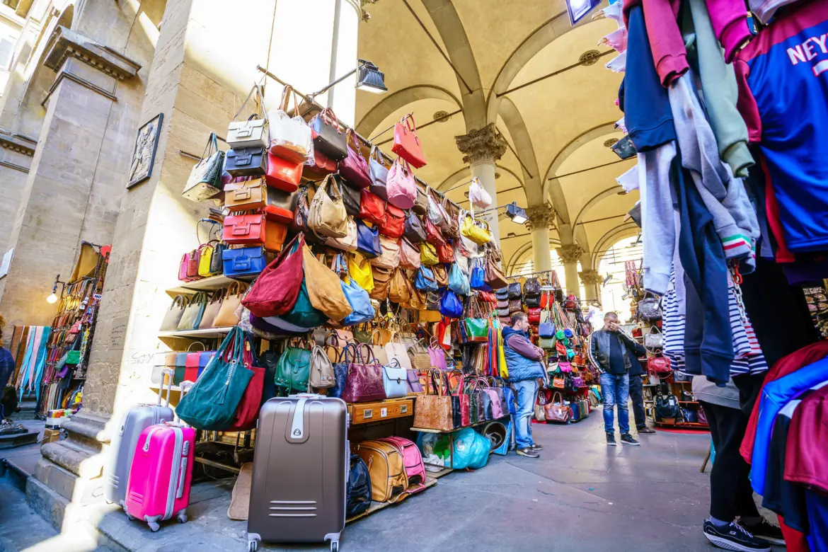 Colorful leather bags, handbags, and goods displayed at market stalls beneath the Renaissance arches of Mercato Nuovo in Florence, Italy