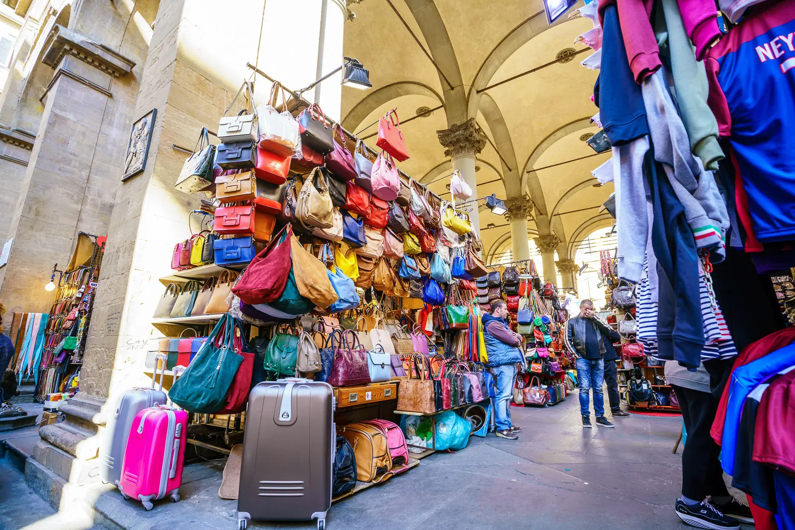 Colorful leather bags, handbags, and goods displayed at market stalls beneath the Renaissance arches of Mercato Nuovo in Florence, Italy