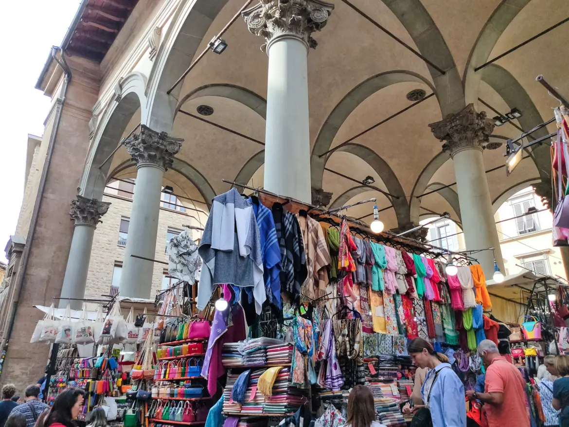 Colorful scarves, shawls, and handcrafted goods hanging beneath the Corinthian columns and vaulted arches of the Mercato Nuovo loggia in Florence, Italy