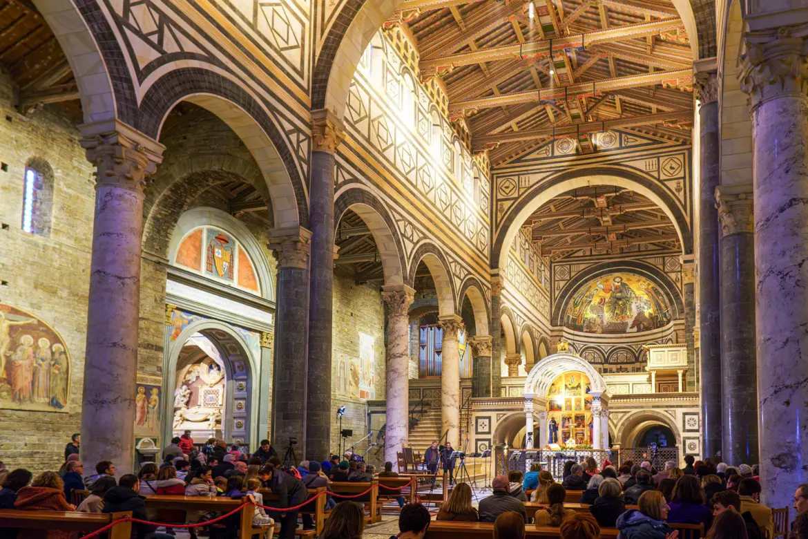 Visitors gathered inside San Miniato al Monte in Florence during a service, with atmospheric lighting illuminating the Romanesque arches, frescoes, and golden apse mosaic