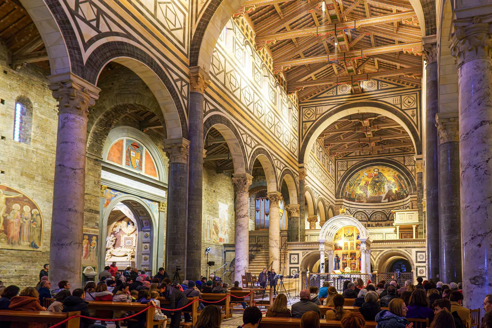 Visitors gathered inside San Miniato al Monte in Florence during a service, with atmospheric lighting illuminating the Romanesque arches, frescoes, and golden apse mosaic