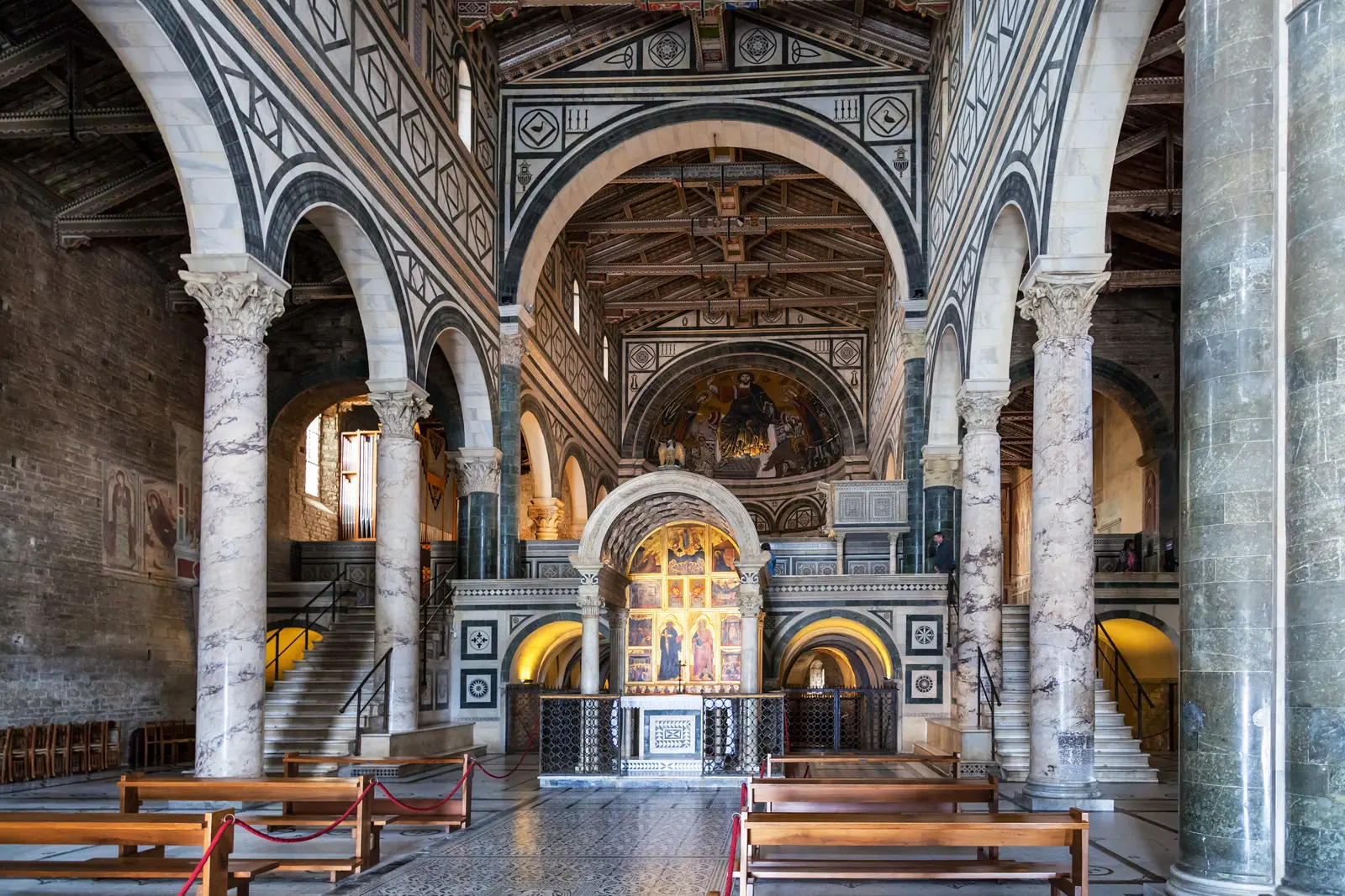 Interior of San Miniato al Monte in Florence showing the Romanesque nave with marble columns, geometric inlaid floor, raised choir above the crypt, golden apse mosaic, and painted timber ceiling