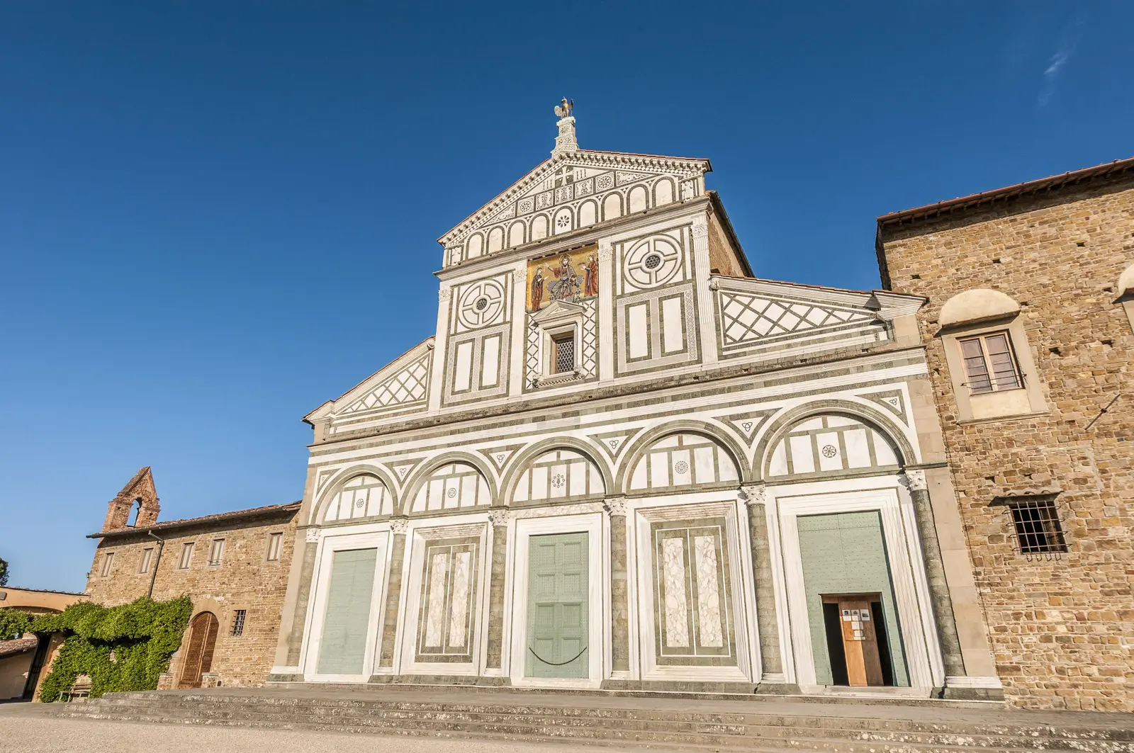 The Romanesque façade of San Miniato al Monte in Florence, with geometric white Carrara and green Prato marble patterns, arched portico, and golden mosaic depicting Christ, the Virgin Mary, and Saint Minias