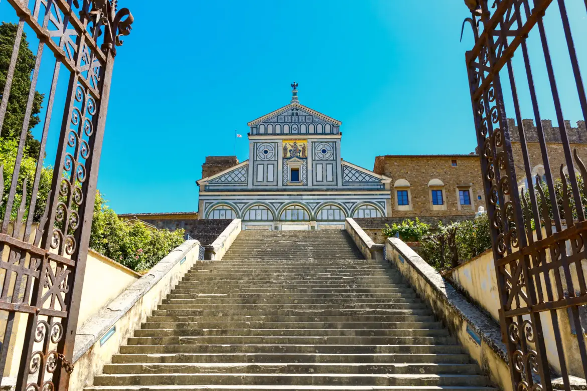 Stone staircase leading to San Miniato al Monte Romanesque church in Florence framed by iron gates with blue sky above