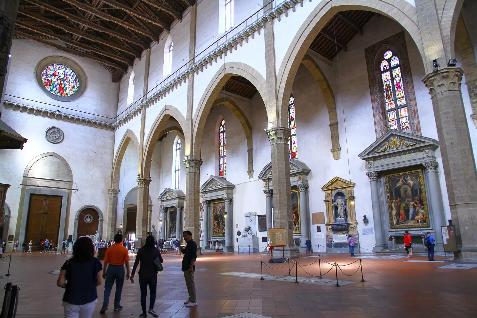 Inside Santa Croce Basilica in Florence with visitors walking through the Gothic nave past side chapels stained glass windows and funerary monuments