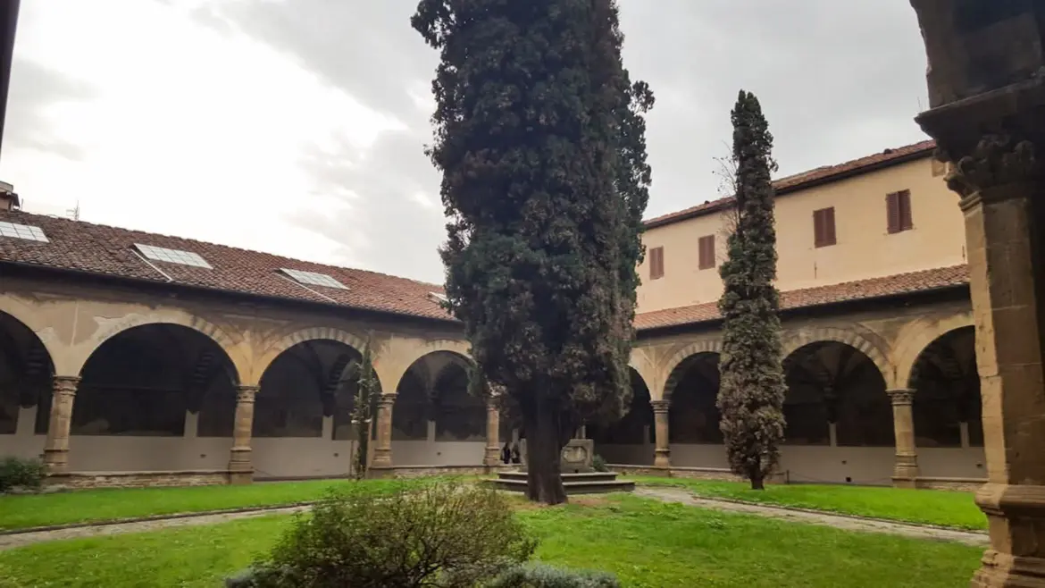 The peaceful cloister of Santa Maria Novella in Florence with graceful stone arcades, cypress trees, green lawn, and overcast sky creating a contemplative atmosphere