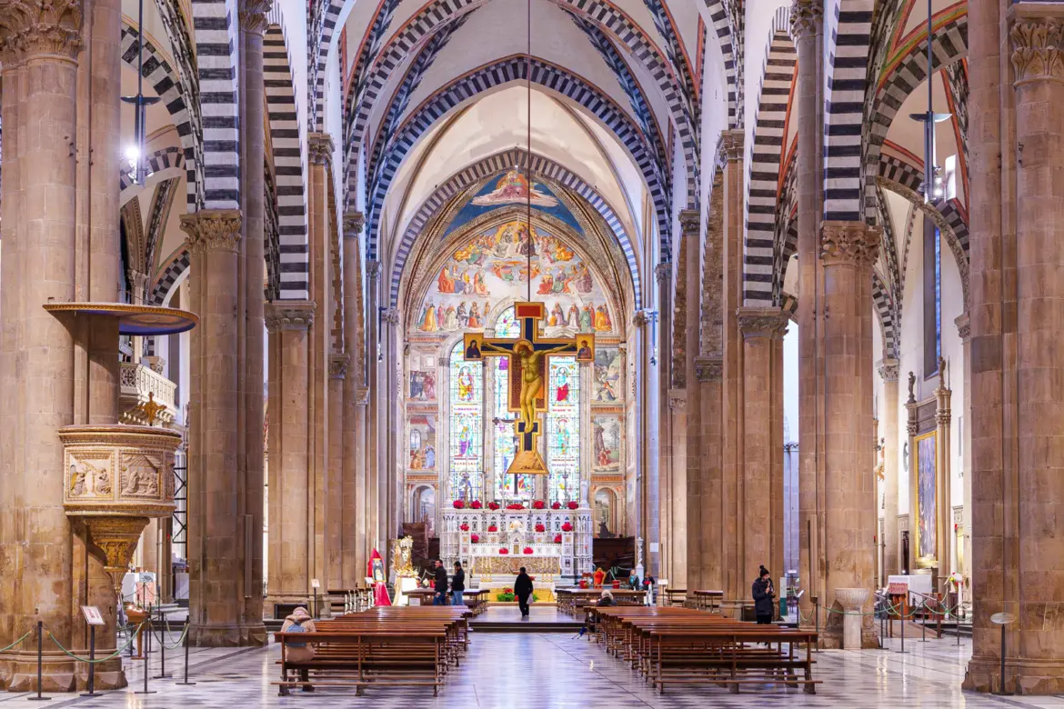 The nave of Santa Maria Novella in Florence looking toward the high altar with Giotto’s painted crucifix suspended above, frescoes by multiple masters, stained glass windows, and the ornate carved pulpit