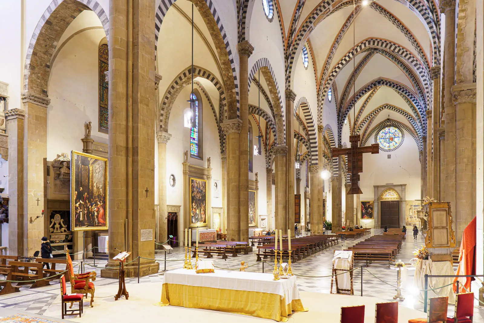 Interior of Santa Maria Novella in Florence viewed from the altar area, showing the long nave with Gothic pointed arches, striped columns, hanging crucifix, wooden pews, and stained glass rose window