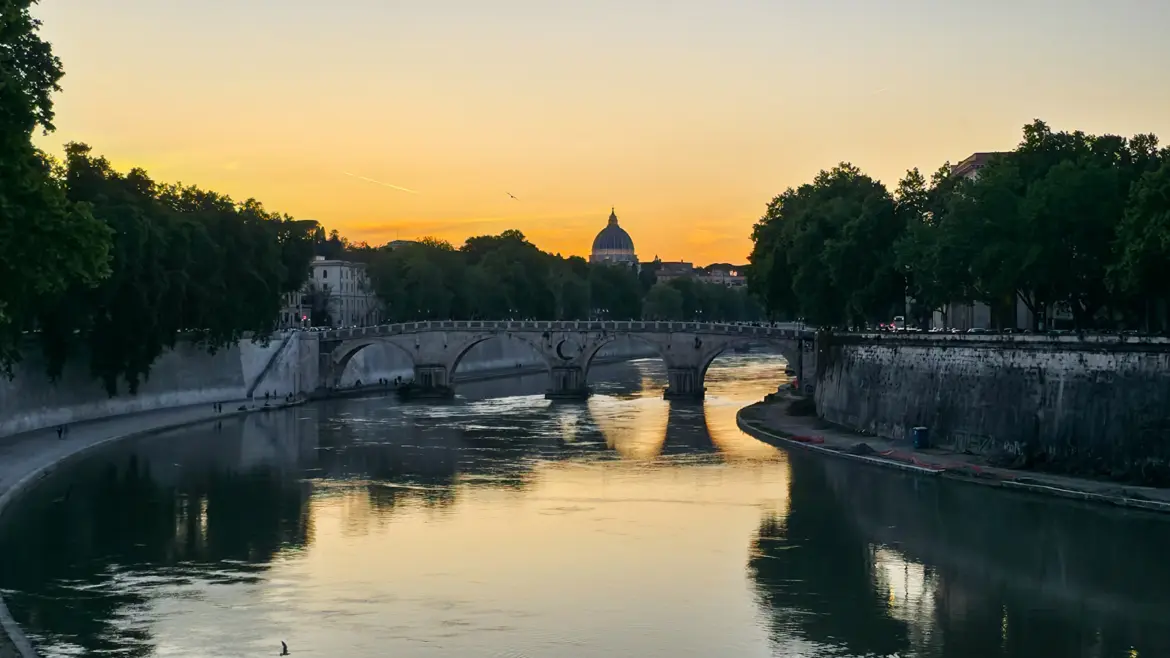 Sunset over the Tiber River in Rome Italy with a historic bridge St Peter’s dome silhouetted in the distance and golden reflections on the water