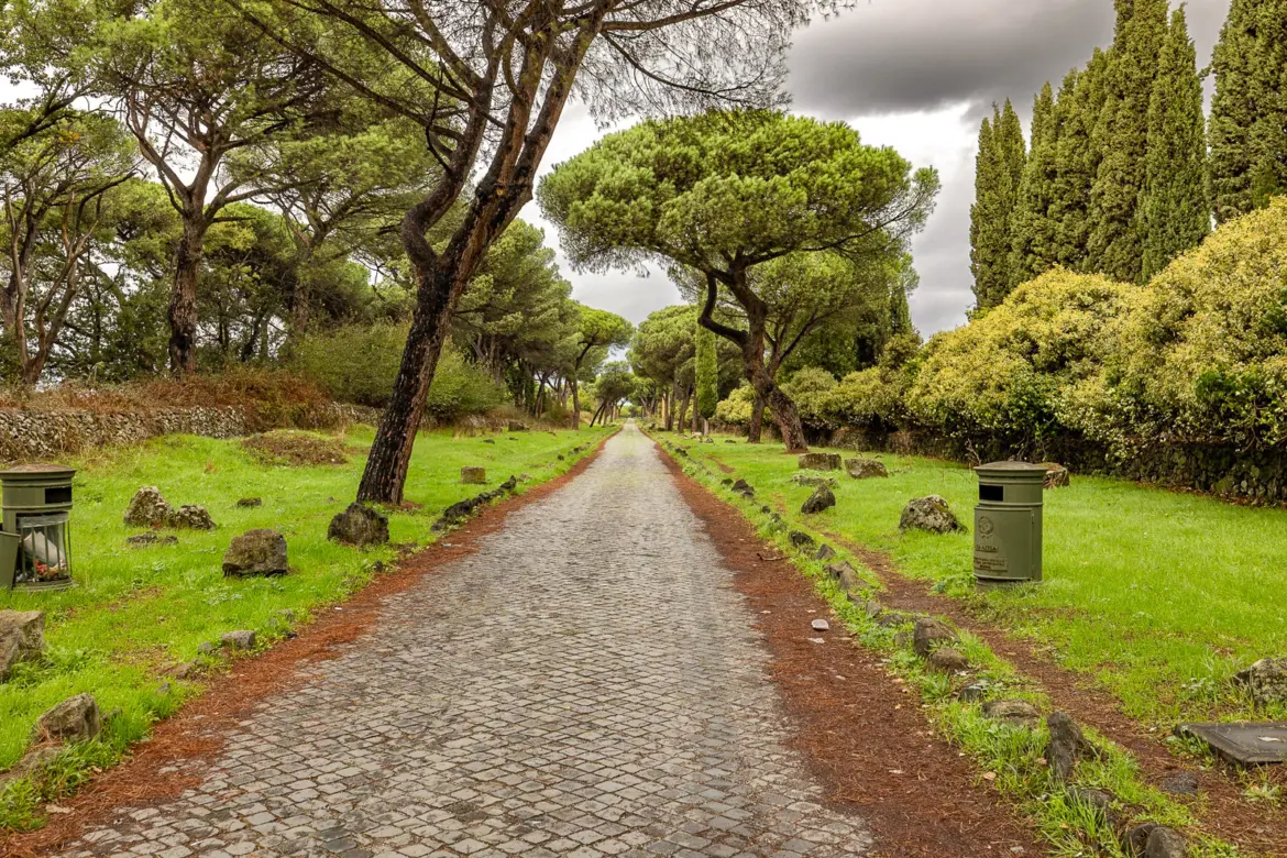 Via Appia Antica ancient Roman road with original paving stones stretching into the distance lined by umbrella pines and greenery under overcast sky