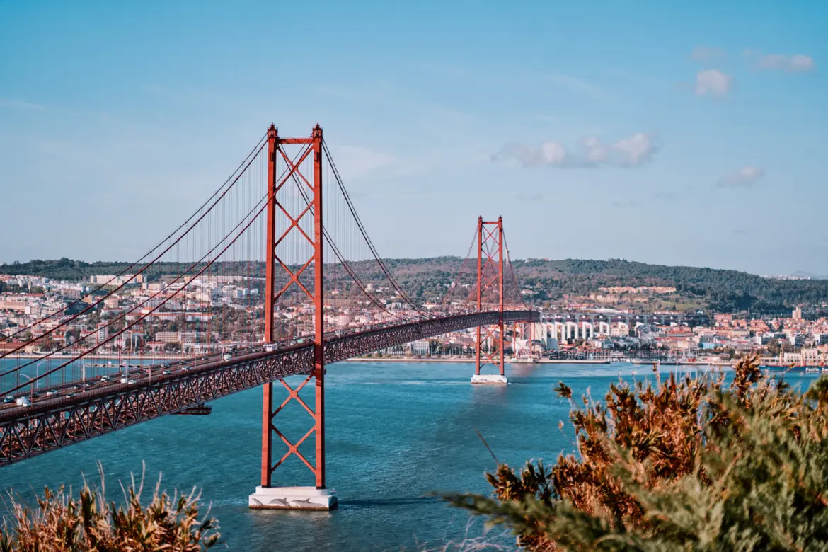 The 25 de Abril Bridge spanning the Tagus River in Lisbon with red suspension towers and Almada hills in the background