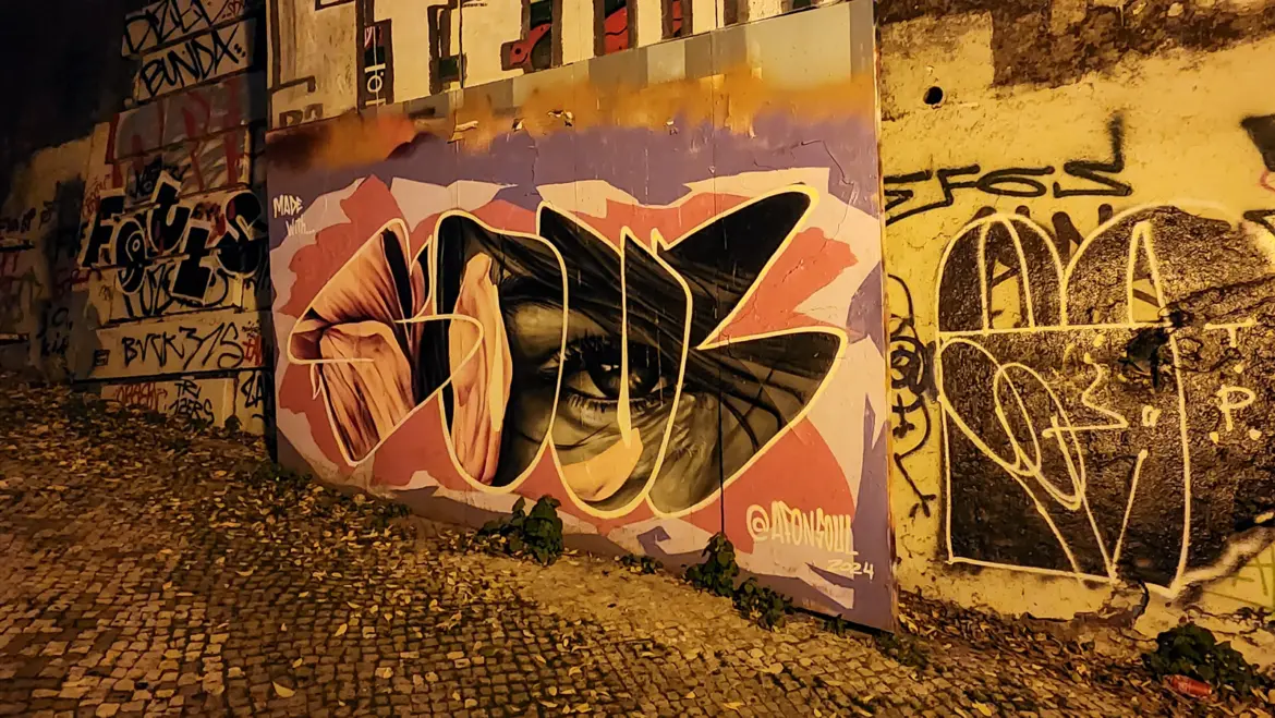 Portrait mural by AtonSoul on a Lisbon street wall at night blending realistic eyes with bold graphic lettering