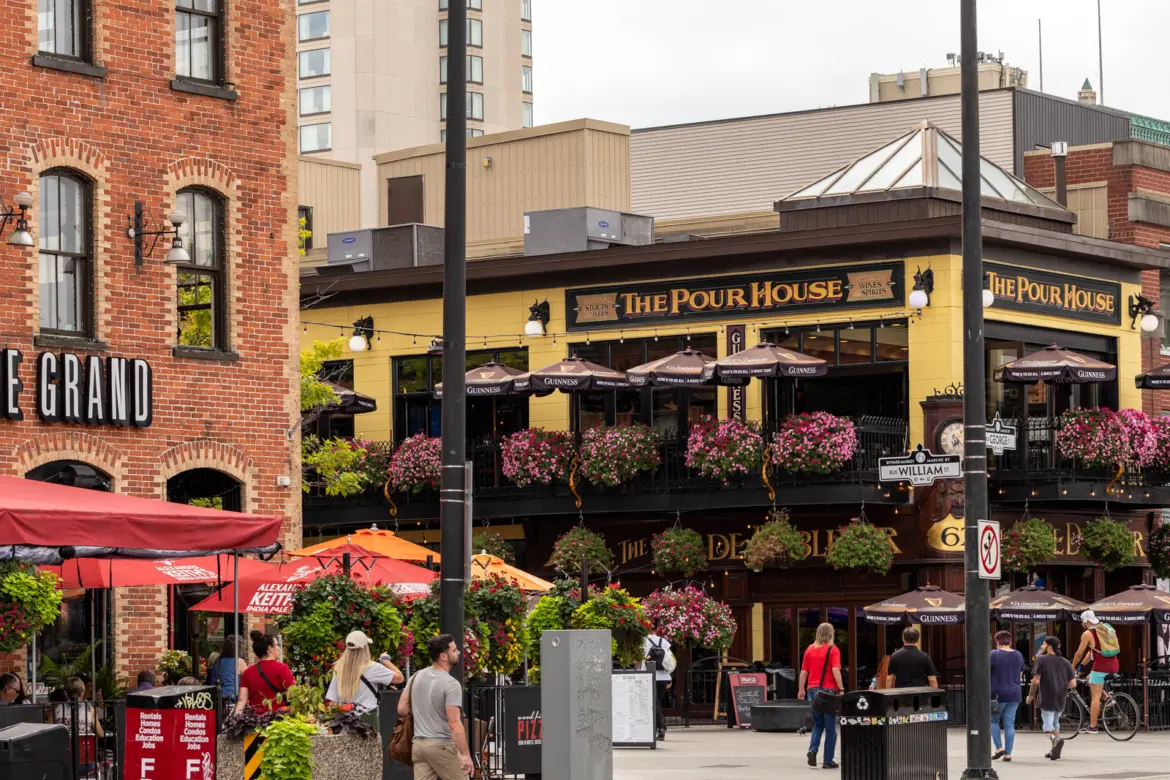 The ByWard Market in Ottawa in summer with The Pour House pub, flower-filled patios, heritage brick buildings, and pedestrians on William Street