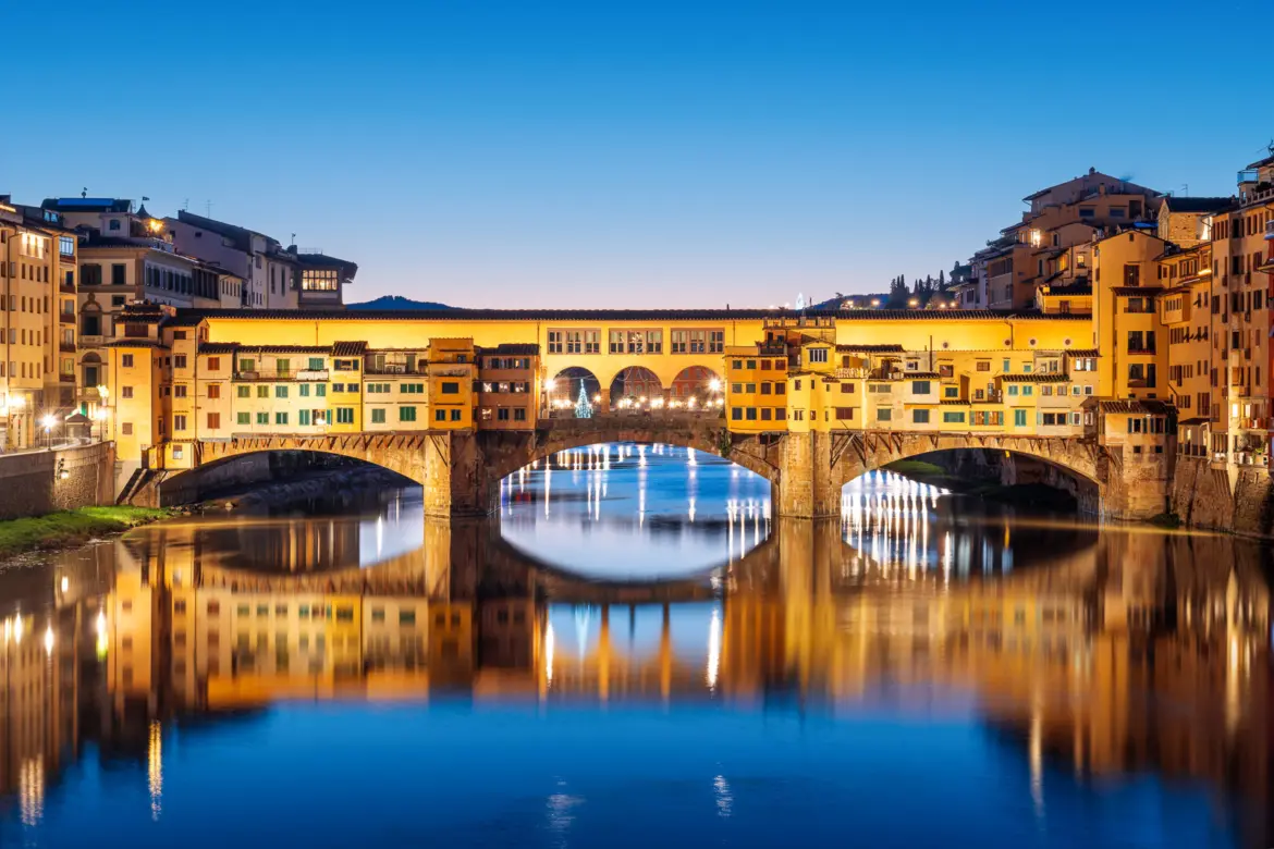 Ponte Vecchio illuminated at night in Florence, reflecting beautifully over the calm Arno River with the Renaissance skyline in the background