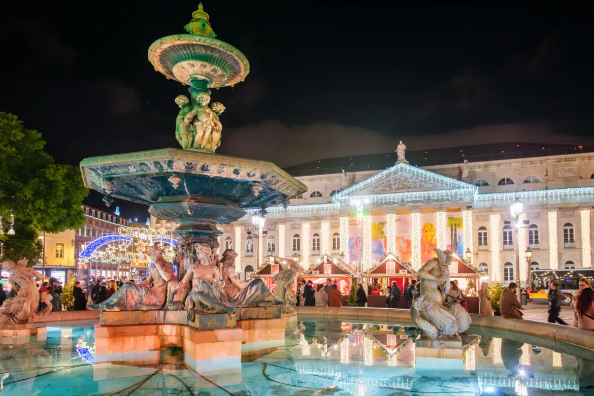 Baroque fountain and illuminated National Theatre D. Maria II at the Rossio Christmas Market in Lisbon at night with wooden chalets and festive lights