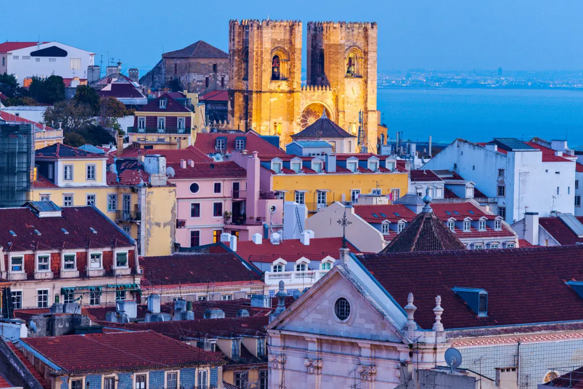 Lisbon Cathedral Sé twin towers rising above Alfama terracotta rooftops with the Tagus River at twilight