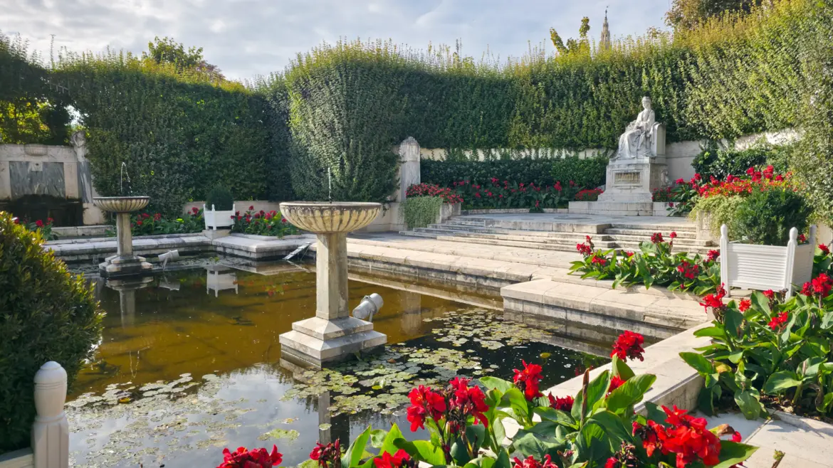 The Empress Elisabeth Monument in Vienna’s Volksgarten with the seated Sisi statue, reflecting basin with lily pads, fountain urns, and red flowers framing the memorial