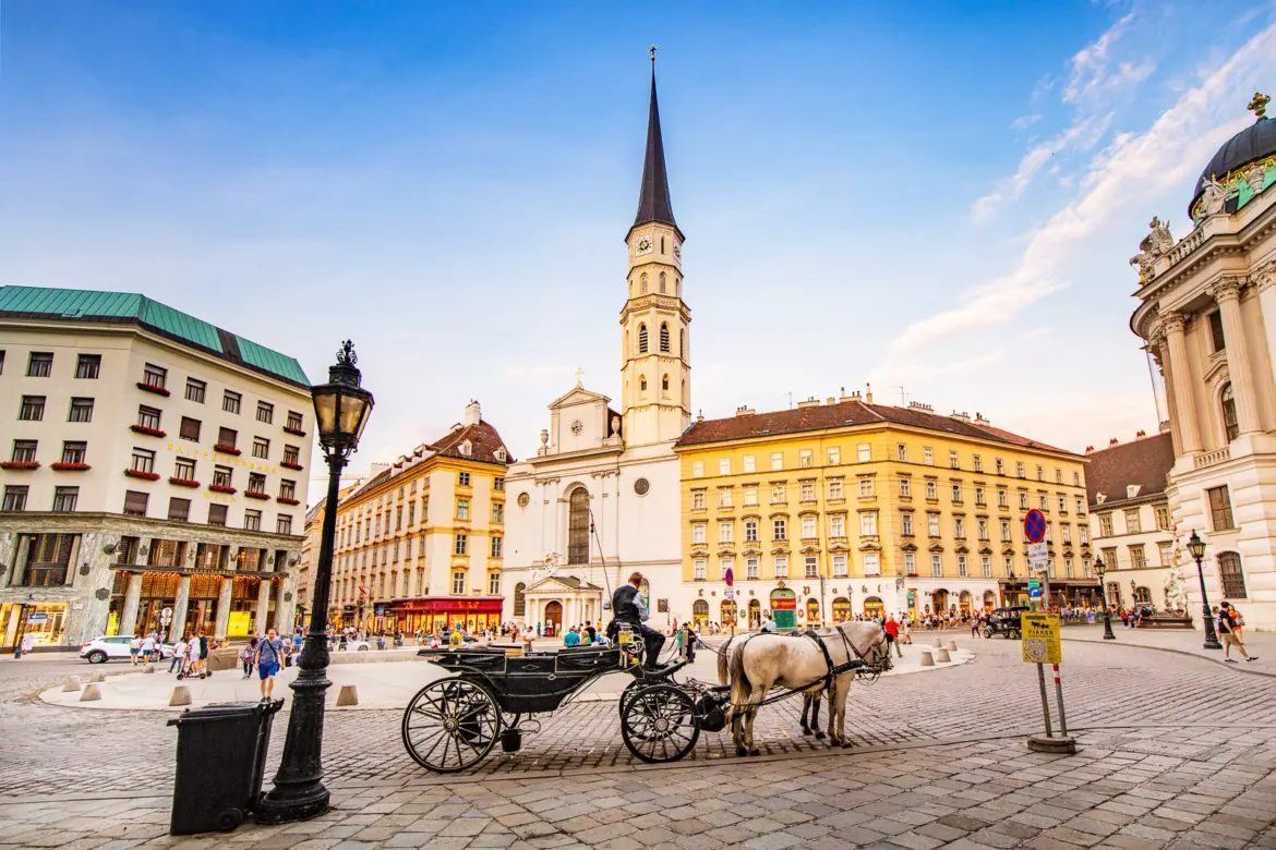 St. Michael’s Church (Michaelerkirche) at Michaelerplatz in Vienna with the Gothic bell tower rising above surrounding buildings and a horse-drawn carriage in the foreground