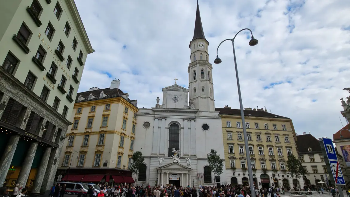 Exterior of St. Michael’s Church at Michaelerplatz in Vienna, showing the Neoclassical façade, Gothic bell tower, and Mattielli sculpture above the entrance