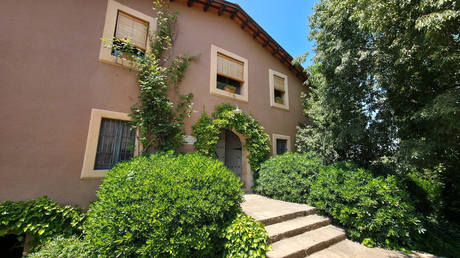 The beautifully preserved Masia Roqueta at Abadal Winery, a medieval farmhouse dating to the 12th century with stone archways, climbing vines, and an arched wooden door framed by lush greenery