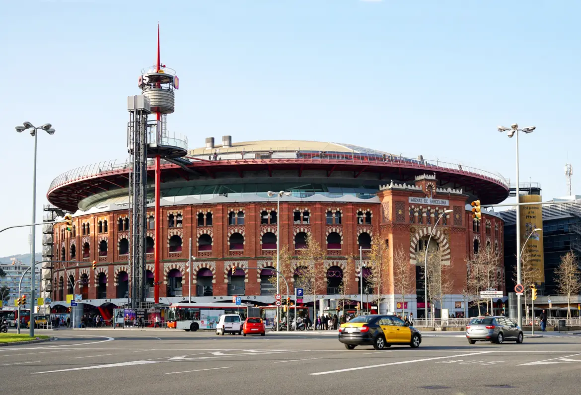 Wide street view of Arenas de Barcelona at Plaça d'Espanya showing the red-brick Neo-Mudéjar bullring façade and Richard Rogers UFO-style modern dome roof with red panoramic elevator tower