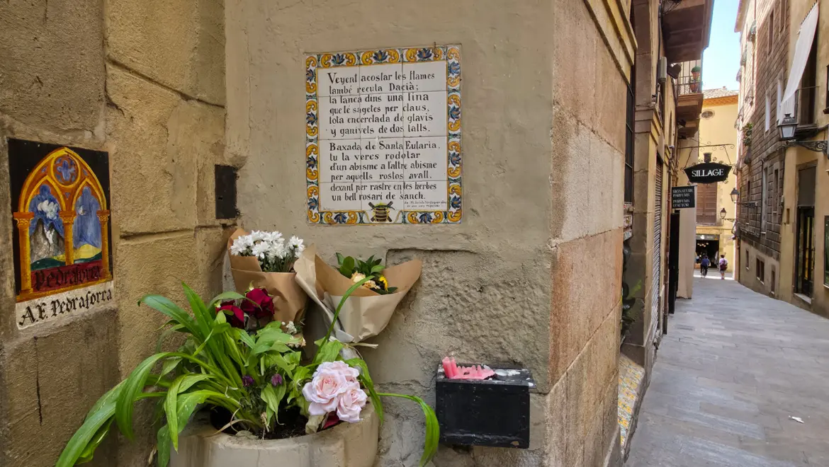 Ceramic tile plaque with Catalan verse about Saint Eulàlia and governor Dacian on the wall of the Baixada de Santa Eulàlia, with fresh flower bouquets and pink candles at the base, Gothic Quarter, Barcelona, Spain
