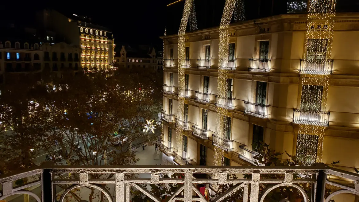 Passeig de Gràcia in Barcelona illuminated with cascading festive Christmas lights viewed from a balcony above, with Modernist facades glowing golden beneath the December night sky