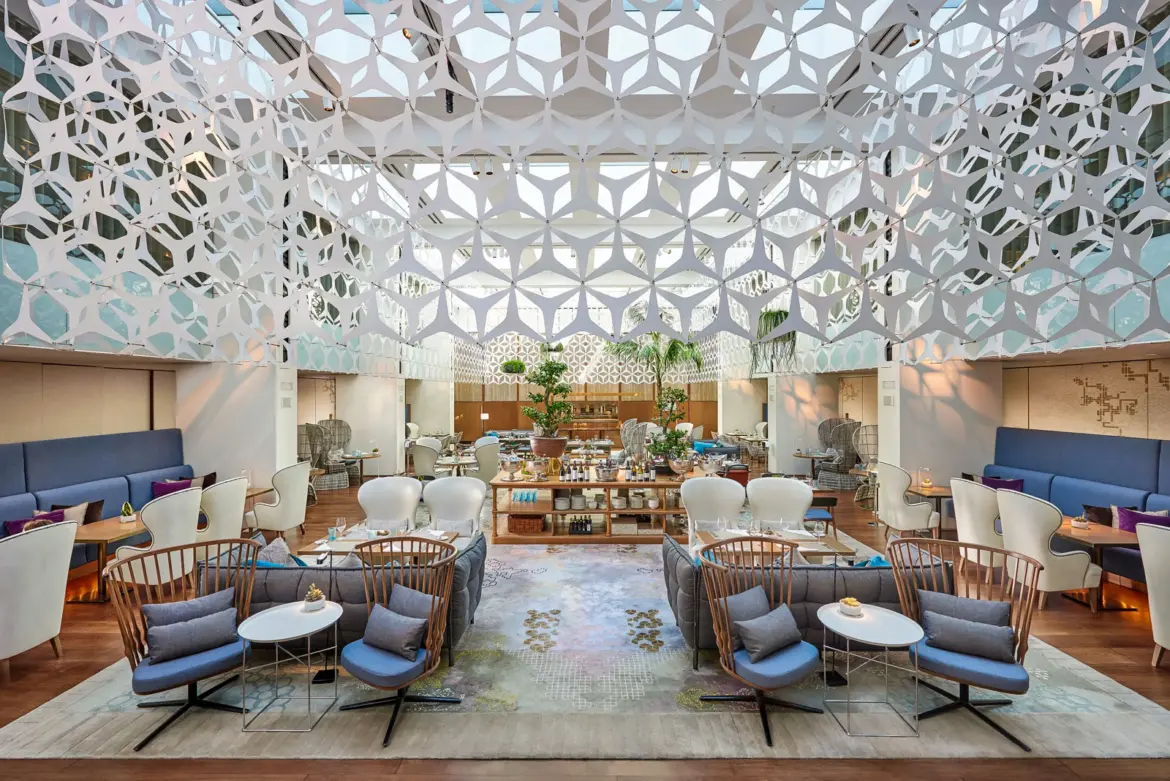 The luminous glass atrium interior of Blanc restaurant at the Mandarin Oriental in Barcelona, with soaring ceilings, white columns, soft cream fabrics, and golden light filtering through the glass canopy