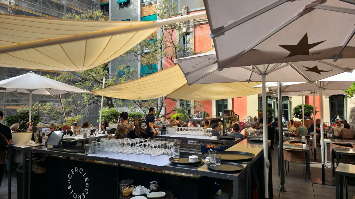 The open-air terrace at El Cercle restaurant in Barcelona's Gothic Quarter, with earth-tone tiles, scattered greenery, and wooden tables glowing under the afternoon sun between Gothic courtyards