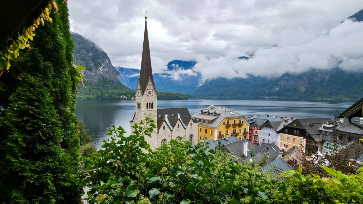 The Evangelische Pfarrkirche Hallstatt viewed from above, with its iconic slender spire rising over the village rooftops and Lake Hallstatt stretching toward the alpine mountains