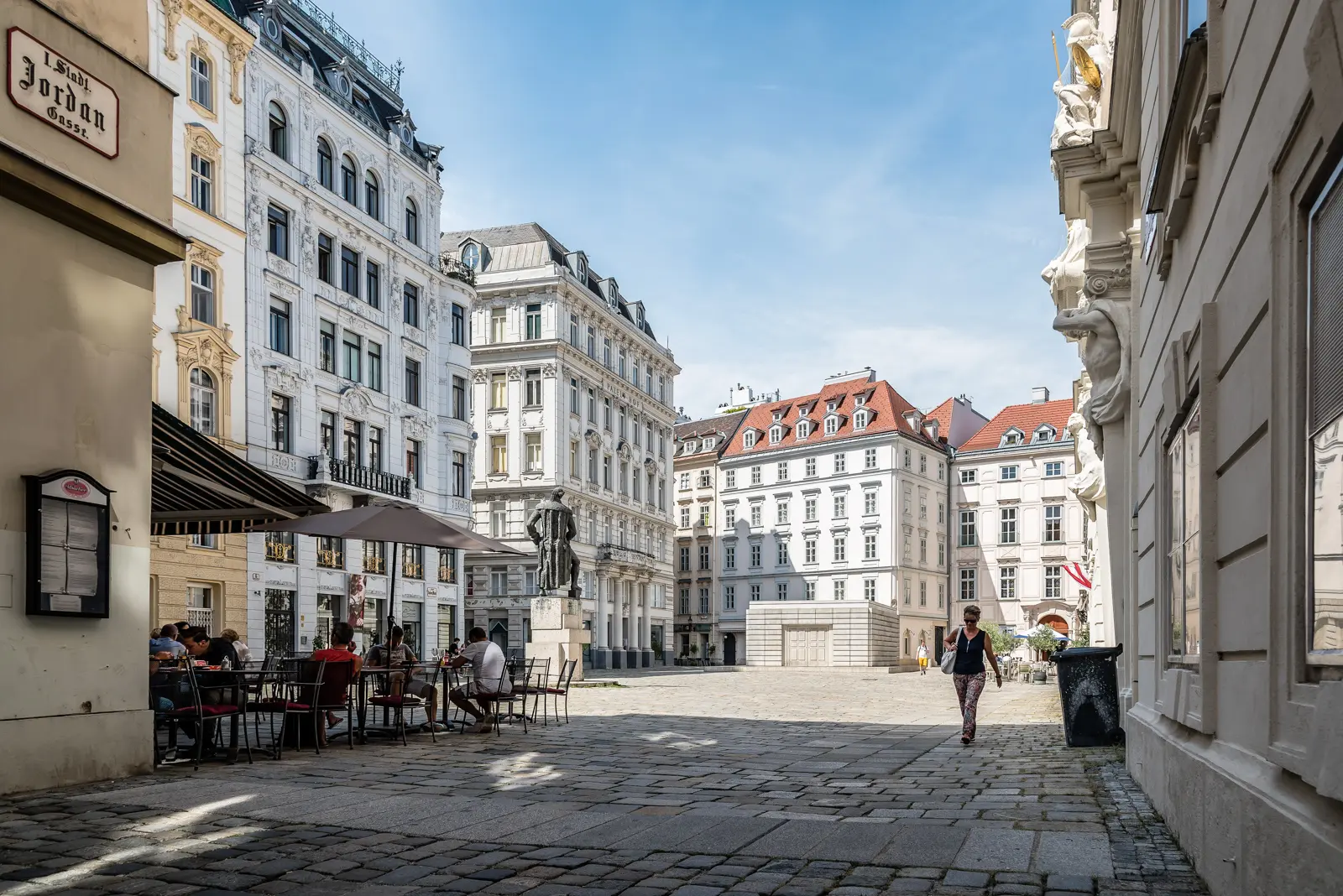 Judenplatz square in Vienna during a sunny afternoon, with cafe tables, cobblestones, the Lessing Monument, and soft natural light shaping the historic facades