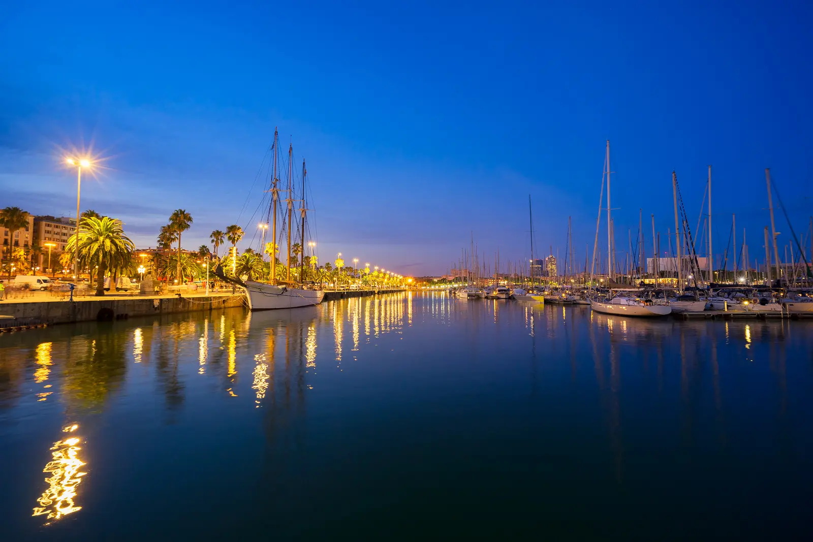 Blue hour at Marina Port Vell Barcelona with streetlight reflections on calm water and a historic tall ship moored along the promenade