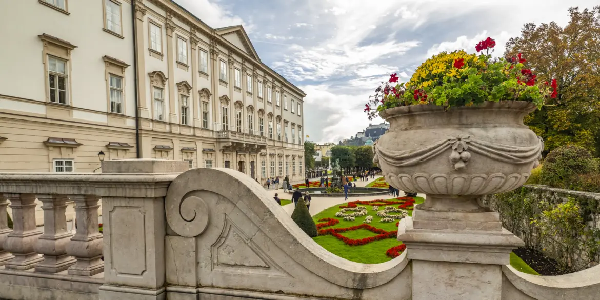 Wide view of Mirabell Palace facade and gardens in Salzburg with a decorative stone urn in the foreground and Hohensalzburg Fortress in the distance
