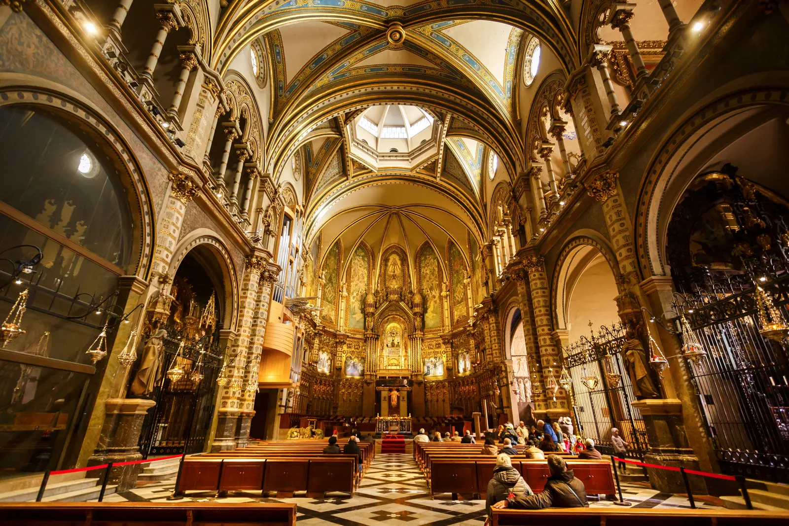 Inside the Basilica of Santa Maria de Montserrat, with tall archways holding pools of quiet light, gold leaf shimmering above the altar, and Gothic lines softened by Renaissance flourishes and warm golden tones