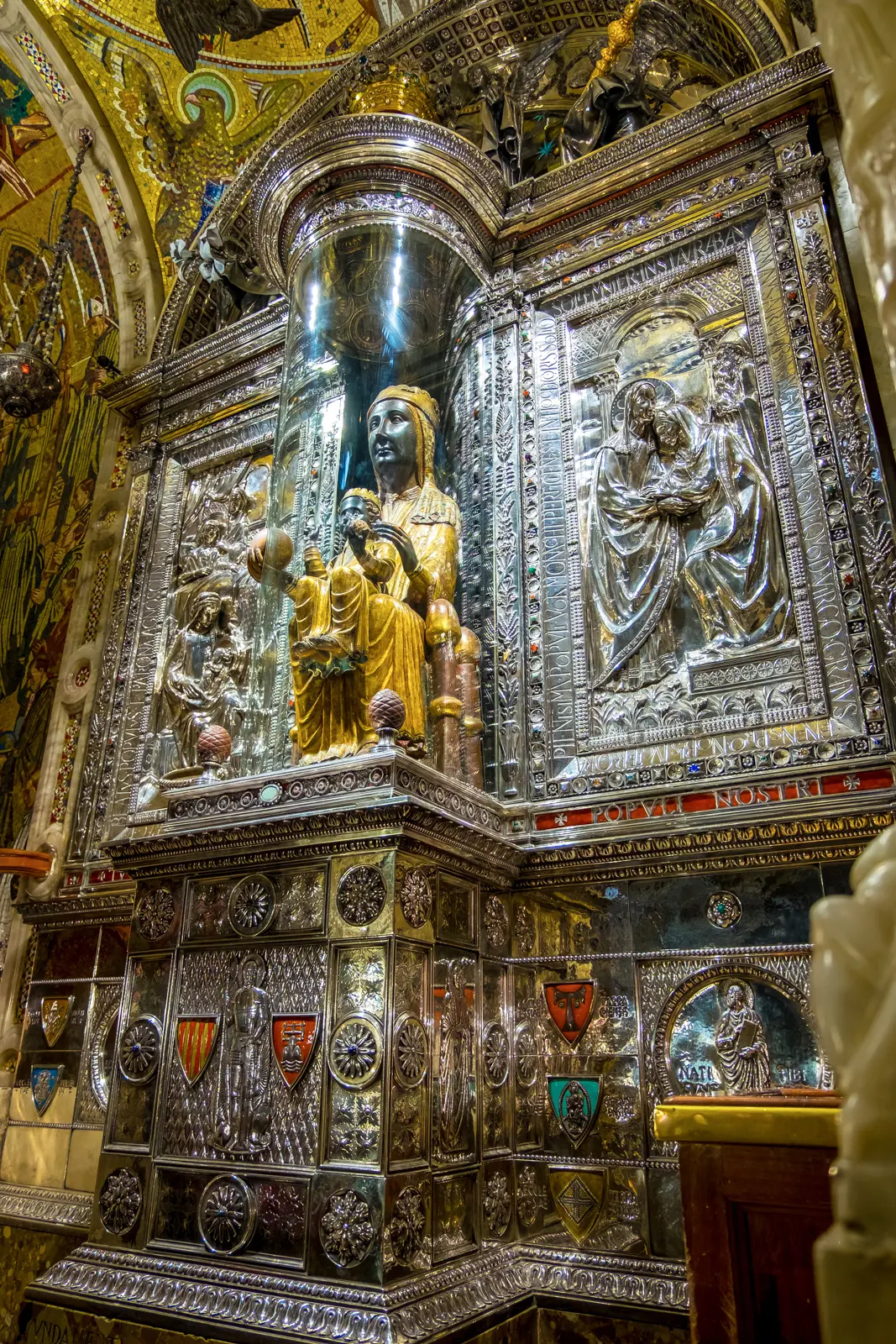 Portrait view of the Black Madonna of Montserrat on her gilded throne, showing the infant Jesus on her lap extending a blessing, surrounded by ornate golden decoration inside the basilica