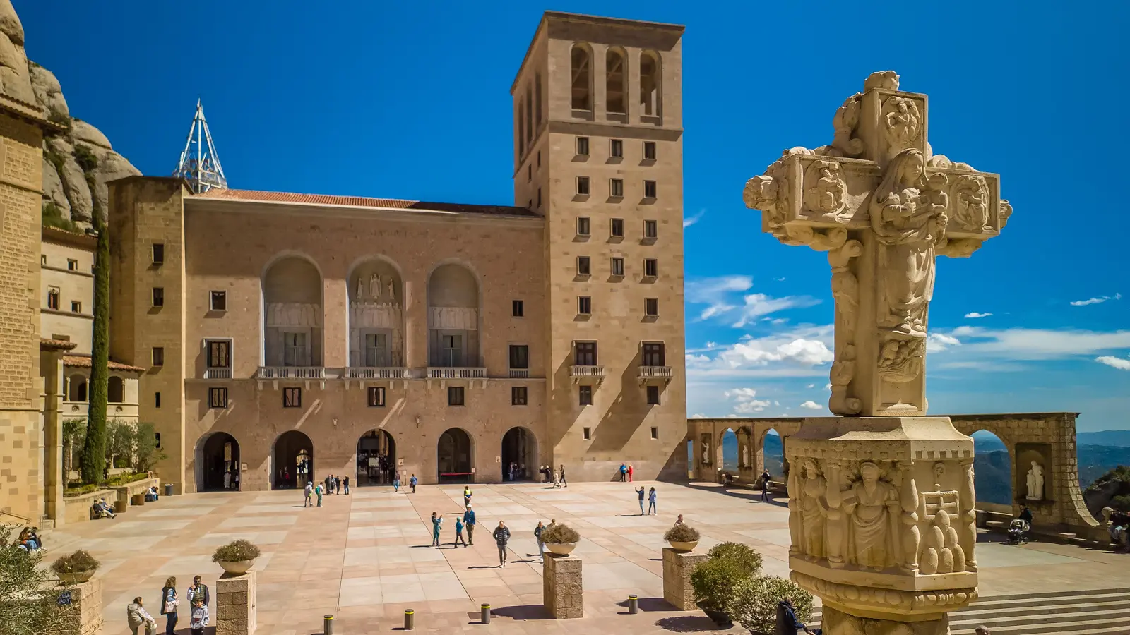 The Benedictine Monastery of Santa Maria de Montserrat seen from a distance, with its ochre buildings nestled against the dramatic serrated mountain peaks under a warm afternoon sky