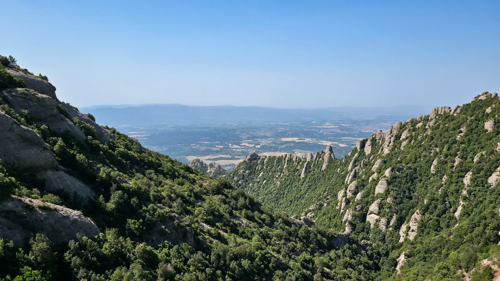 The monastery of Montserrat seen from the Sant Joan summit trail, tiny and golden against the jagged cliffs, looking like a painting set into the mountain's folds far below
