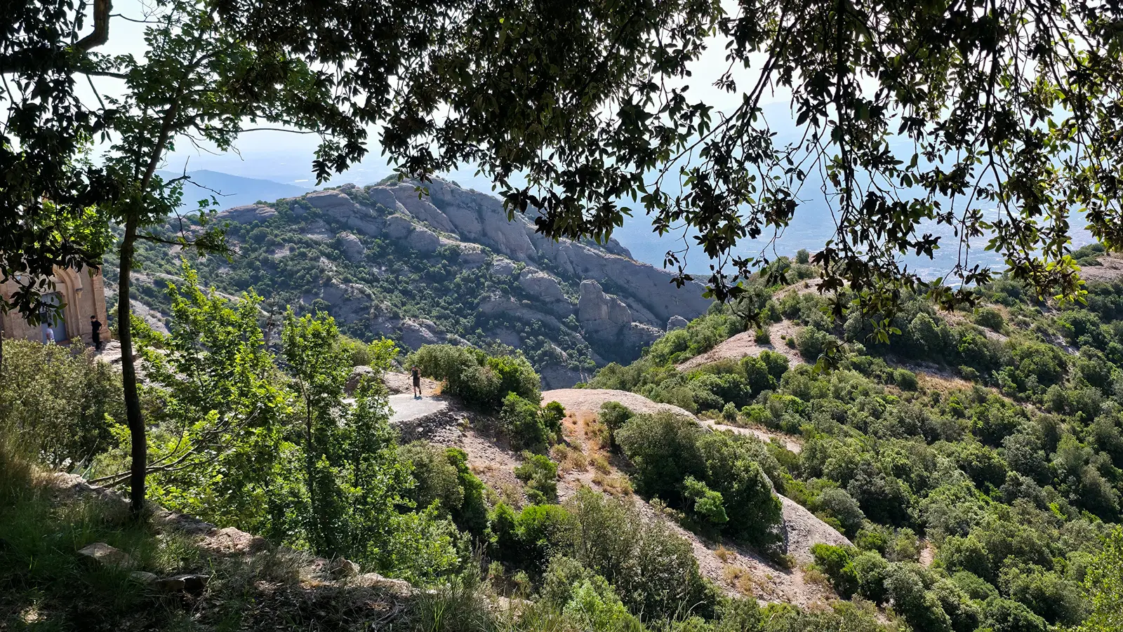 Sweeping panorama from the Sant Joan summit at Pla de les Taràntules, Montserrat, with jagged cliffs, rolling forests, and vast countryside stretching toward Manresa and beyond into a soft blue haze