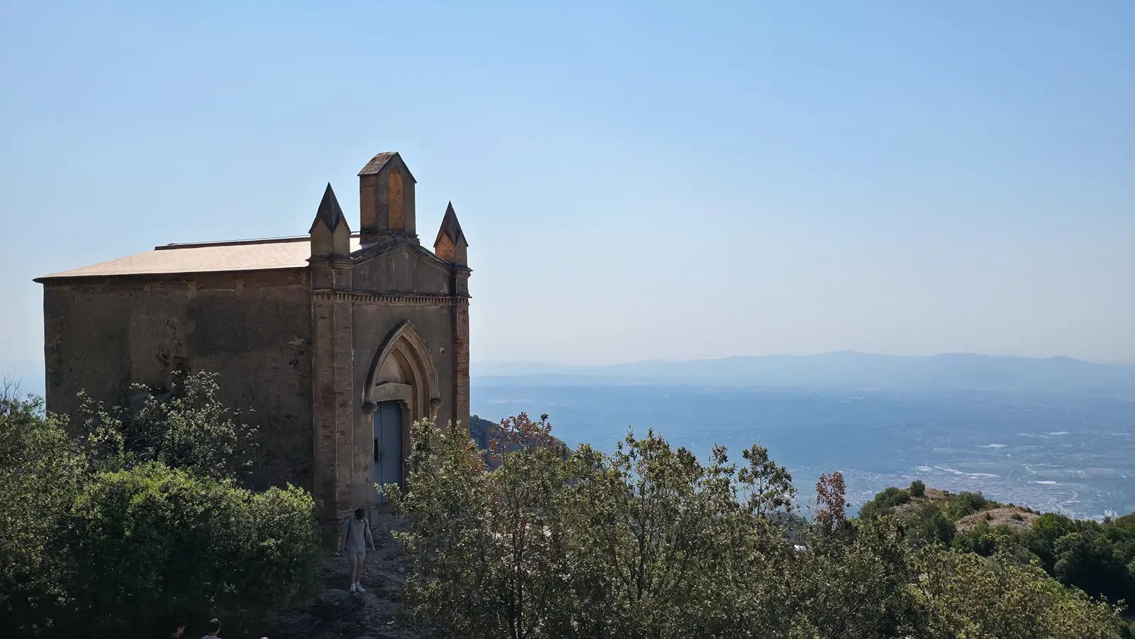 A viewpoint at the Sant Joan summit on Montserrat showing the vast Catalan landscape below, with distant ranges fading into soft blue haze and the monastery tucked into the mountain's folds
