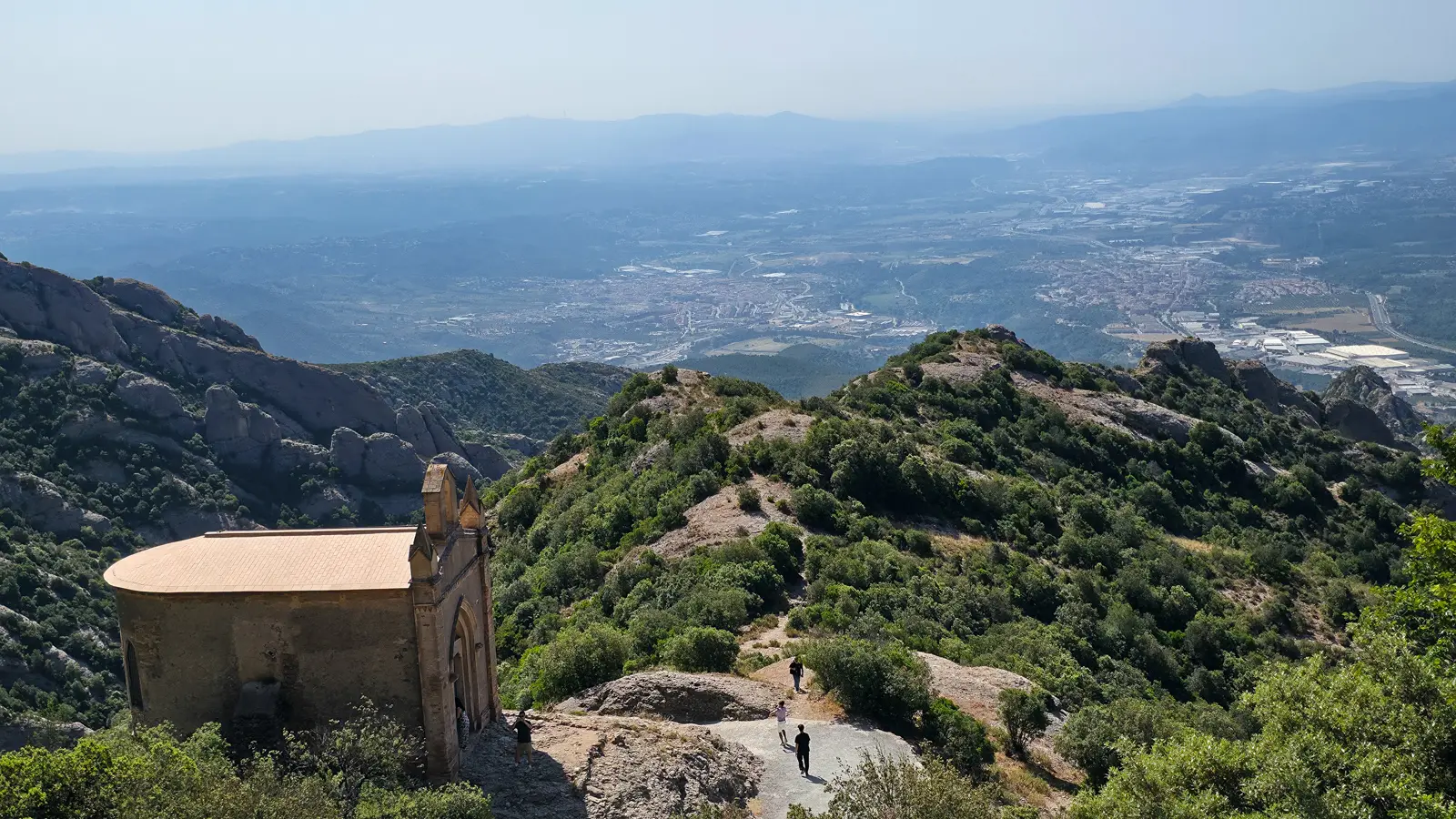 Panoramic view from the Montserrat summit overlooking the Catalan countryside, with layered valleys, forested hills, and the distant horizon melting into a soft blue haze under a warm afternoon sky