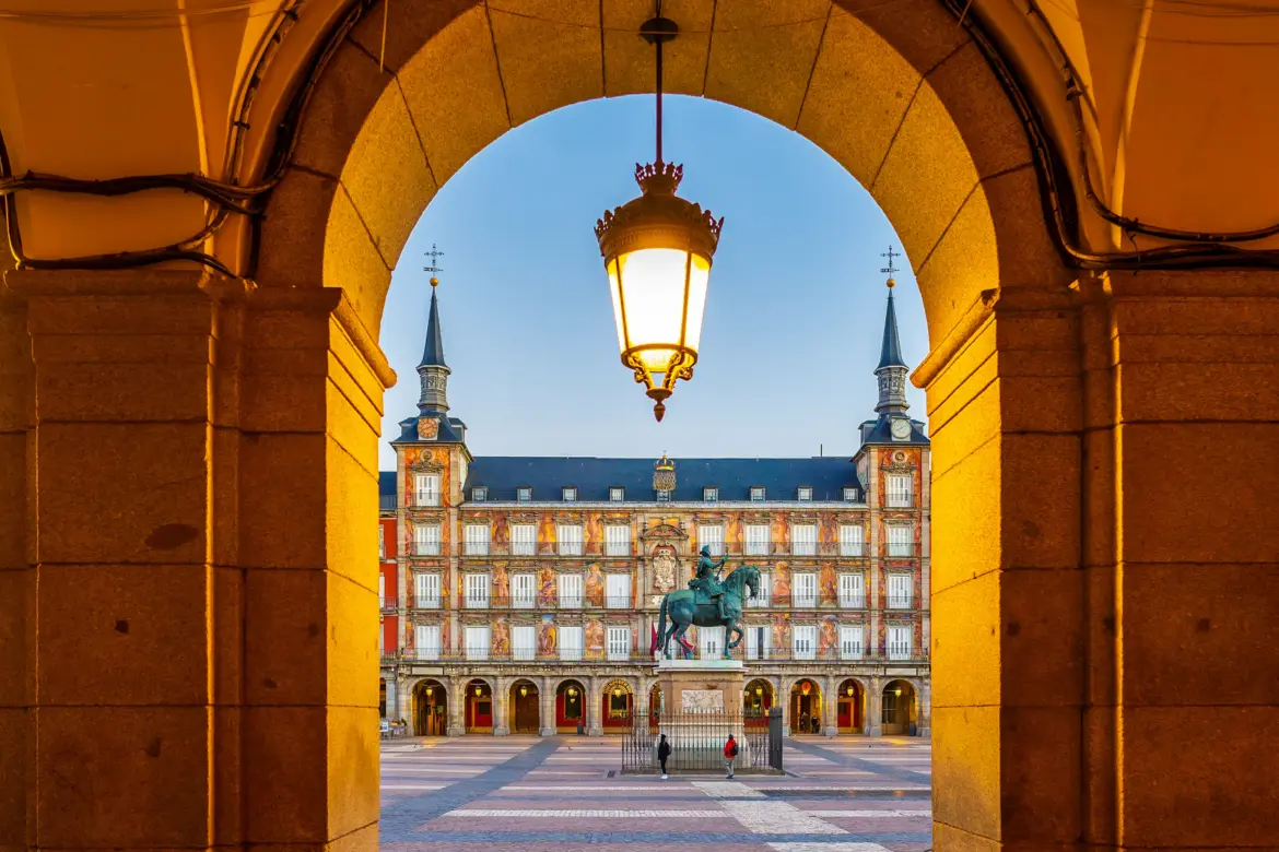 Plaza Mayor in Madrid viewed through a stone arch with an ornate lantern, framing the Casa de la Panadería and the Philip III statue at golden hour