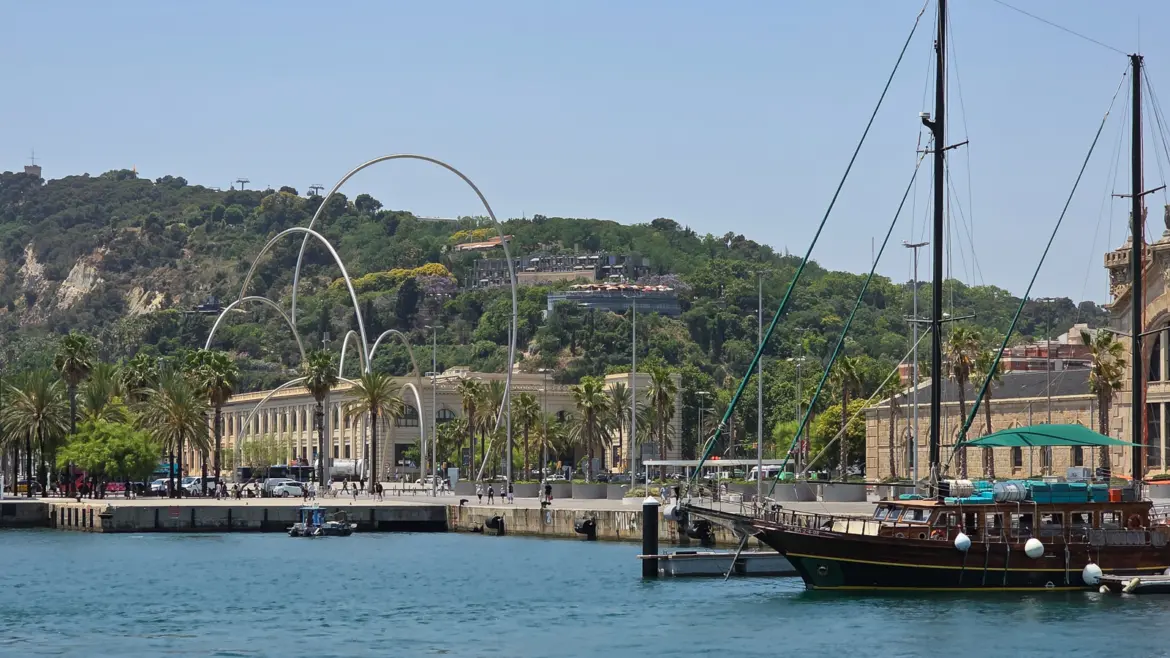 Wide view of Port Vell Barcelona showing Andreu Alfaro Ones sculpture four arched white metal waves Moll de la Fusta promenade Montjuic hill and a historic schooner