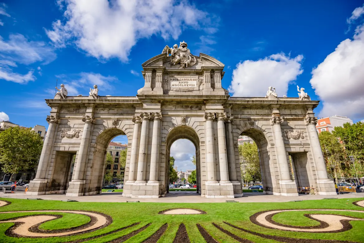 The Puerta de Alcalá in Madrid, a Neoclassical triumphal gate with five arches and sculpted Colmenar stone, set against a blue sky with decorative gardens